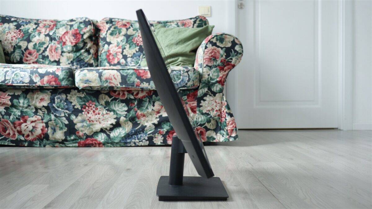 Ecran de calculator pe suport de birou, poziționat în fața unei canapele cu tapet floral, într-un li.